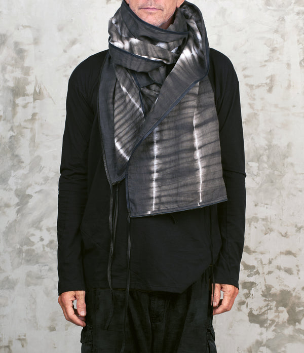Person wearing a black outfit with a gray tie-dye scarf against a textured white wall.